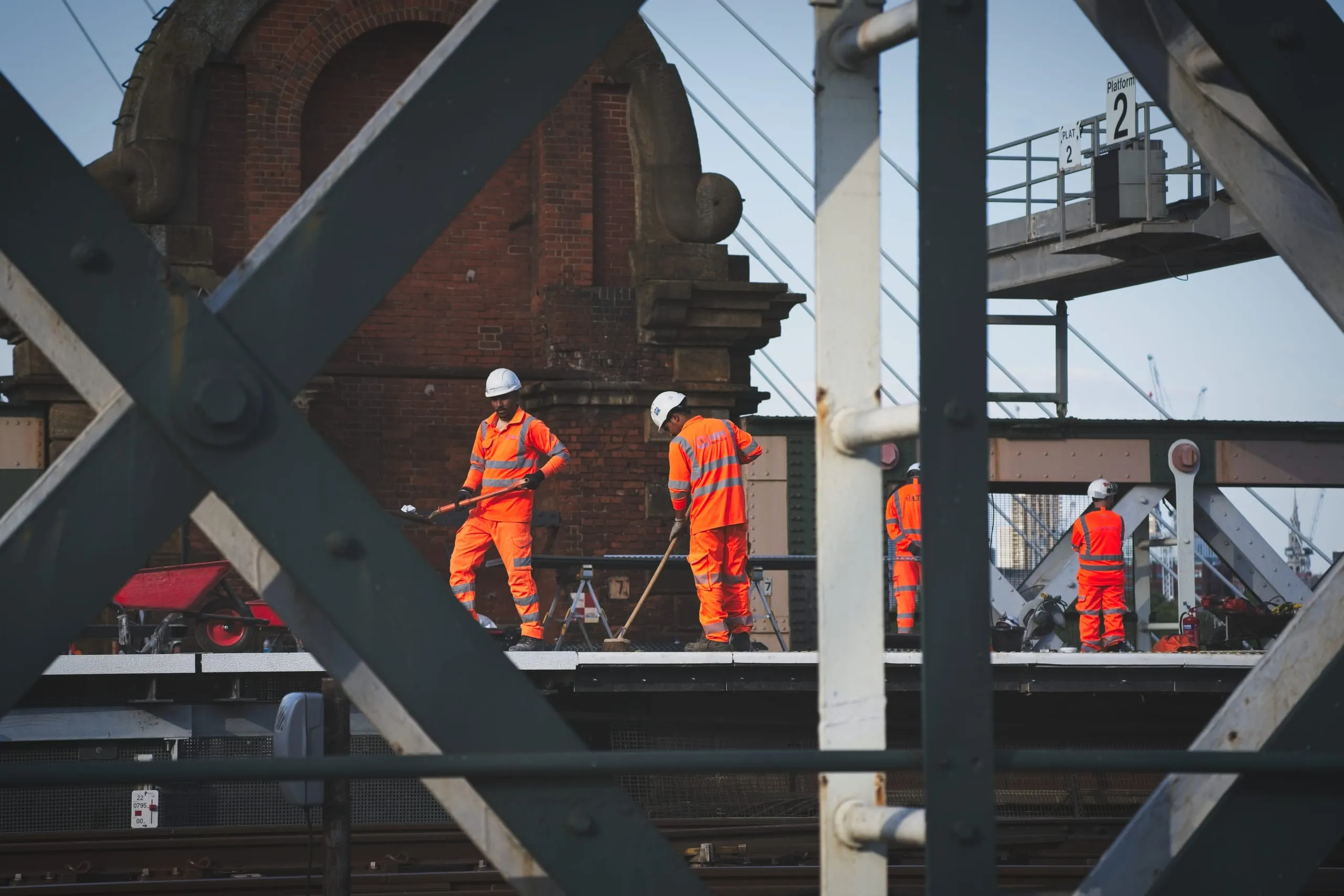 A photograph of railway maintenance workers in orange high-visibility clothing sweeping a platform, framed through steel bridge girders at a London train station.