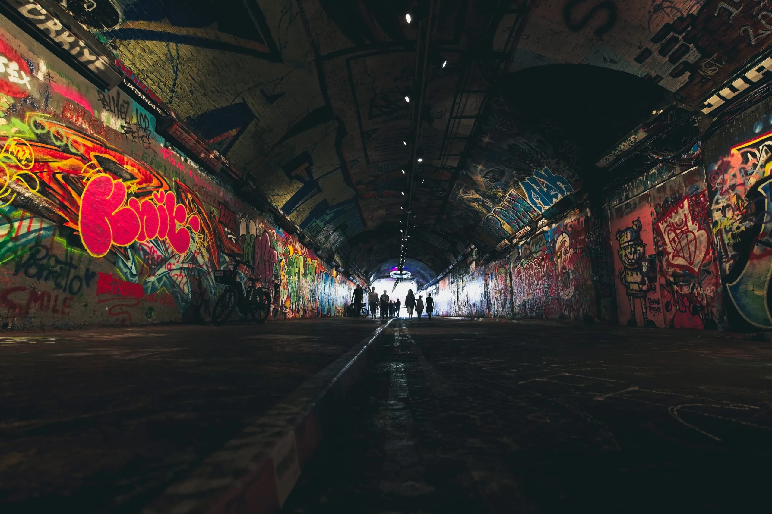 Photograph of a graffiti-covered tunnel in London shot from low and wide, a bicycle leaning against the wall and a small group of people walking toward the bright opening at the far end.