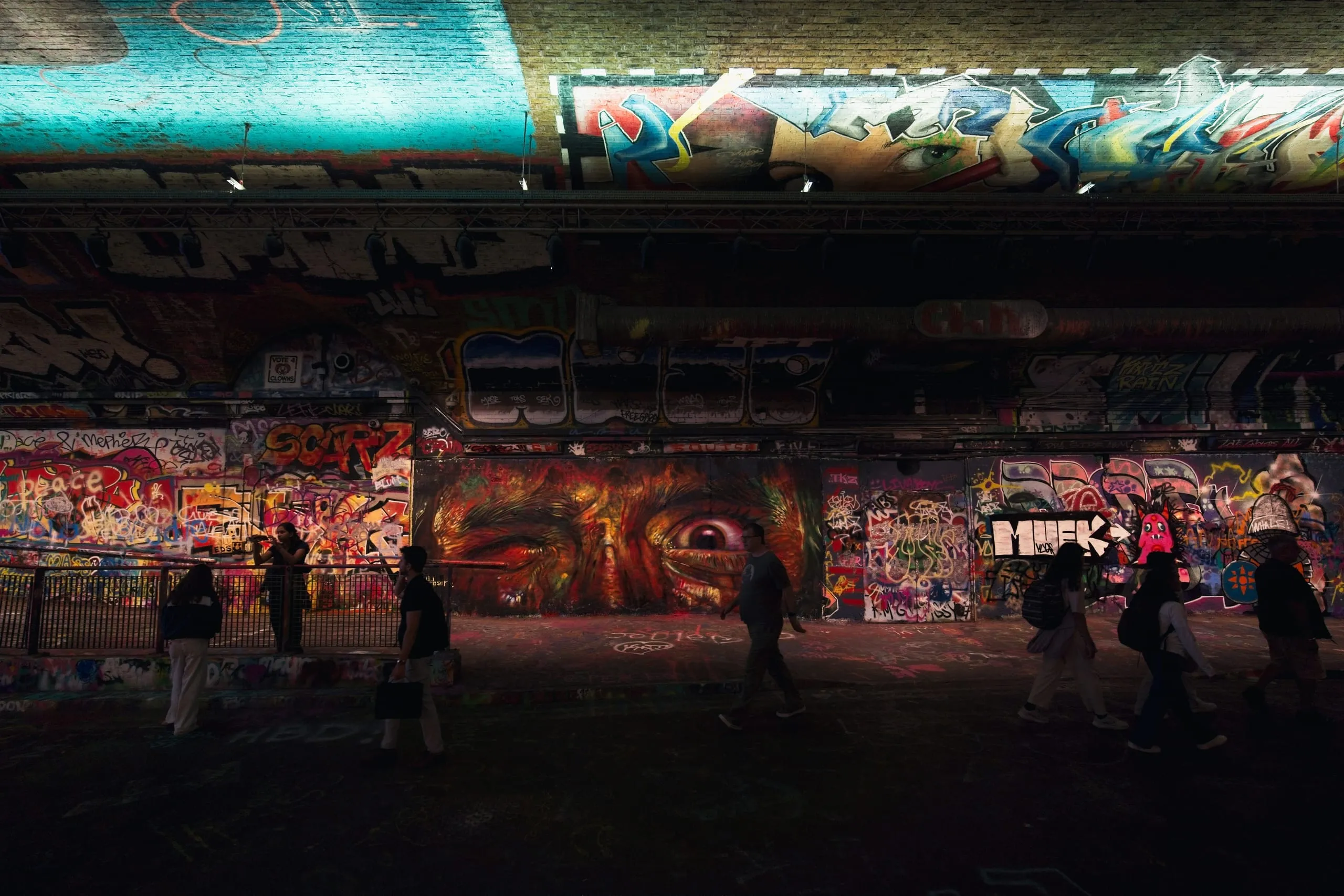 Photograph of pedestrians silhouetted as they walk through the graffiti-covered Leake Street tunnel in London, past a large mural of a fiery-colored face with one eye.