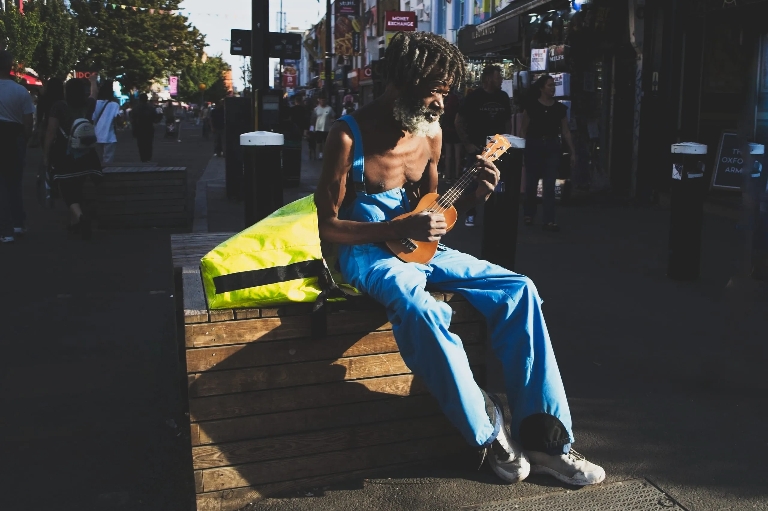 A photograph of a street musician in blue overalls playing a ukulele while seated on a wooden bench along a busy shopping street in Camden, London, England.