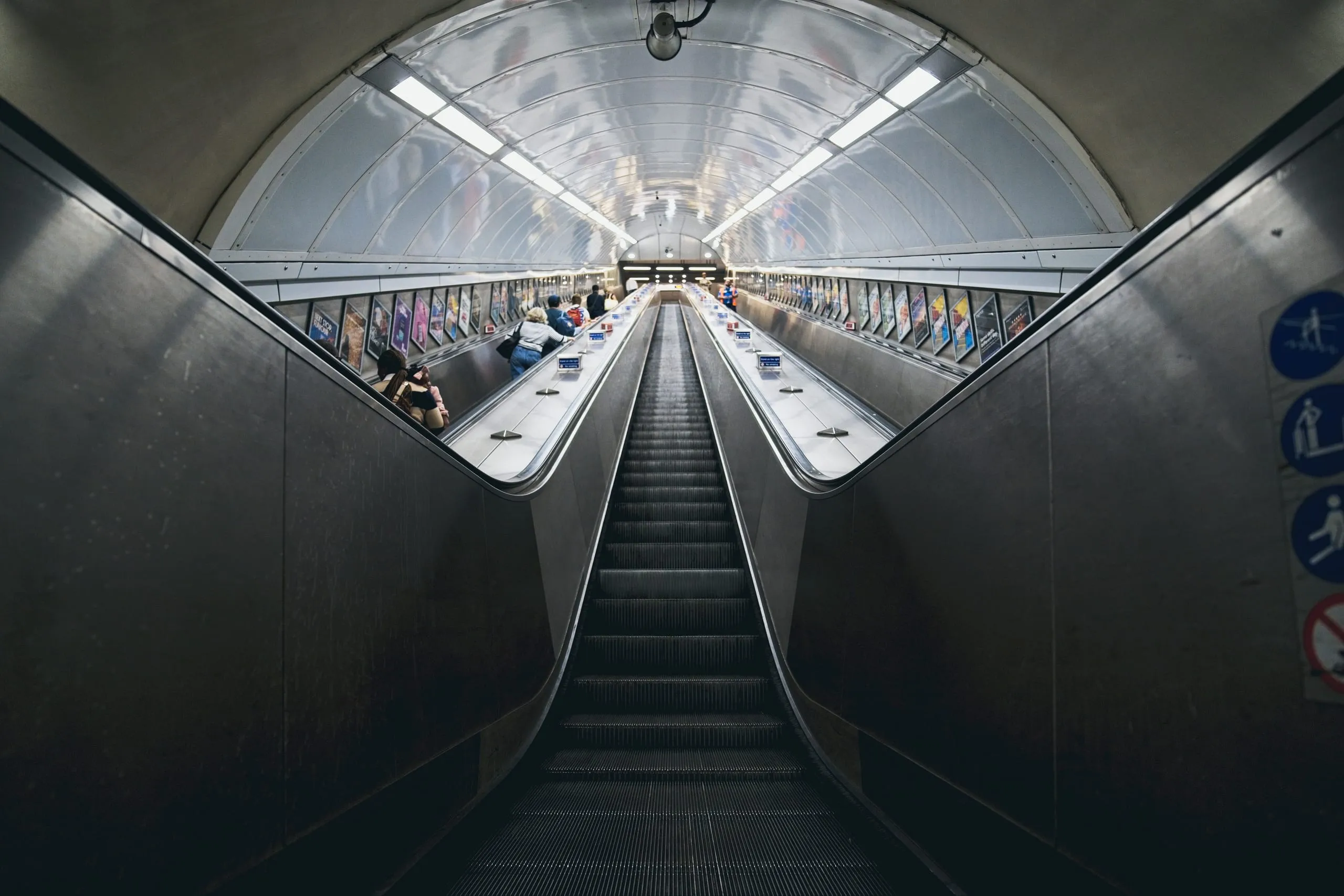 Photograph of a symmetrical London Underground escalator tunnel with illuminated advertising panels lining both walls and passengers riding the opposite escalator upward.