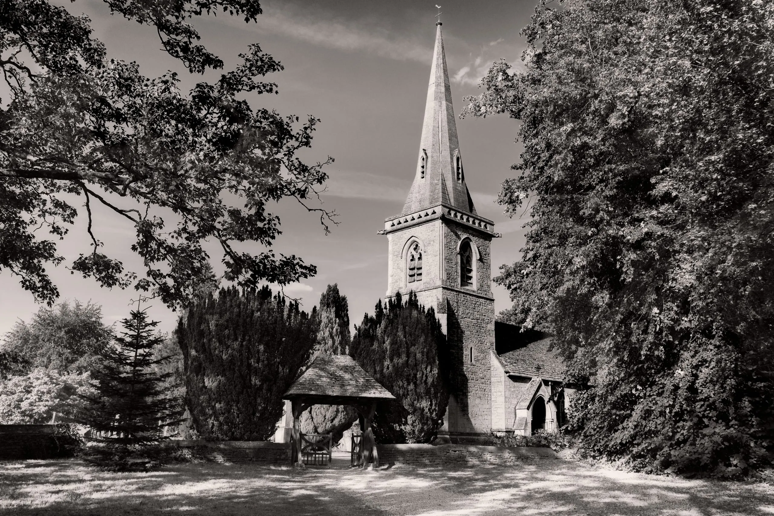 Black and white photograph of a stone village church with a tall spire and a wooden lychgate, framed by trees, in the Cotswolds, England.