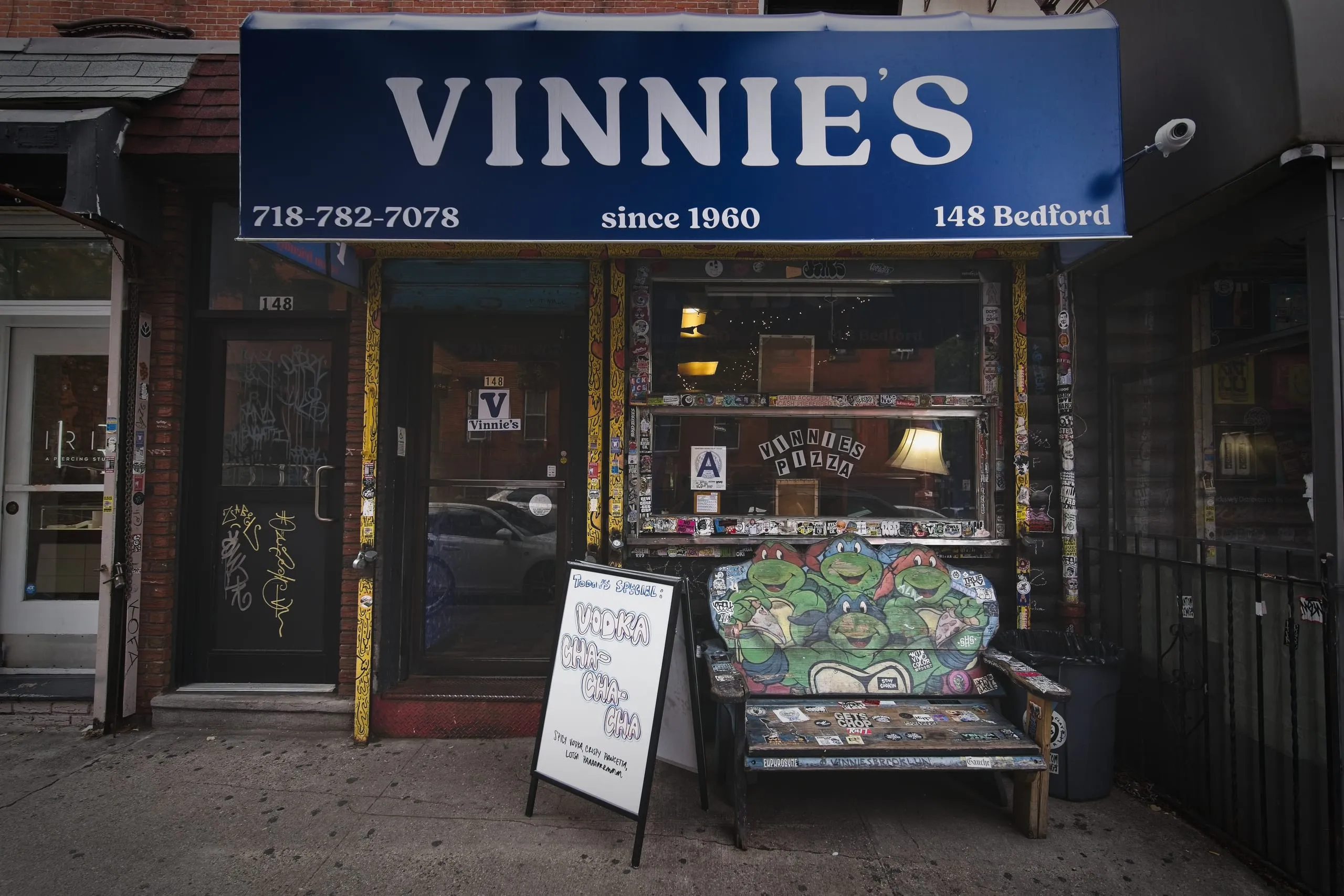A color photograph of Vinnie's Pizza, a Williamsburg, Brooklyn pizzeria storefront with a blue awning reading 'Vinnie's since 1960' and a bench painted with Teenage Mutant Ninja Turtles out front.