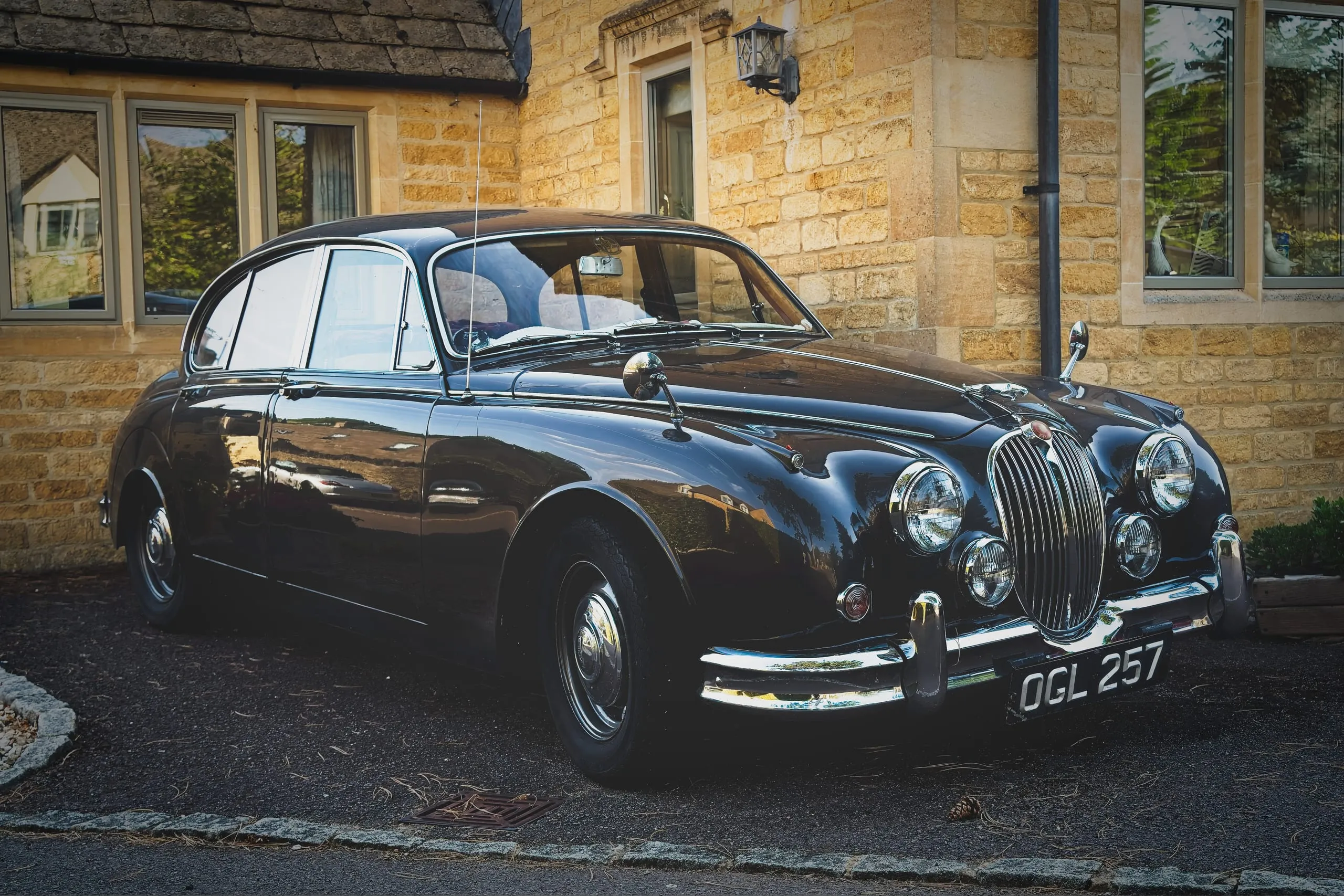 A photograph of a polished vintage black Jaguar saloon parked outside a stone building in the Cotswolds, England.
