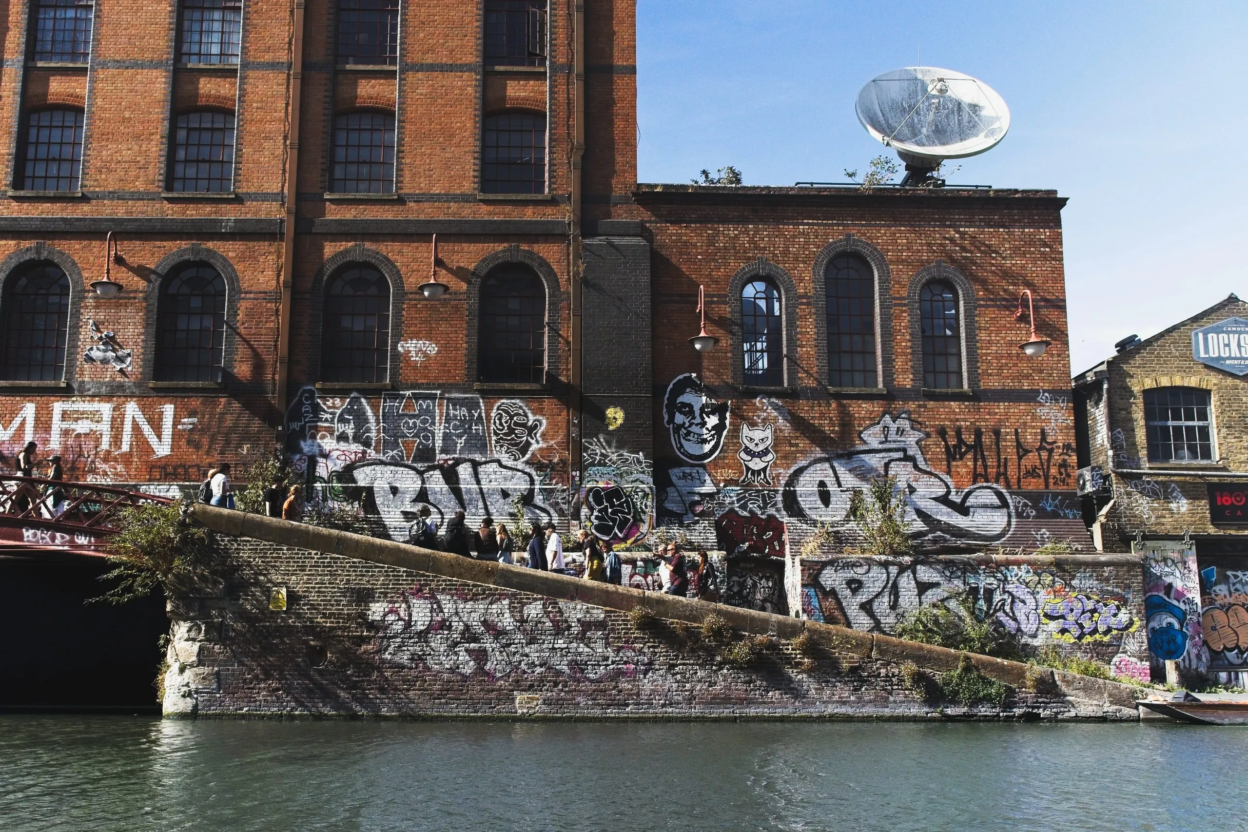 Color photograph of a graffiti-covered brick warehouse with arched windows and a large satellite dish on the roof, along Regent's Canal near Camden Lock, London.