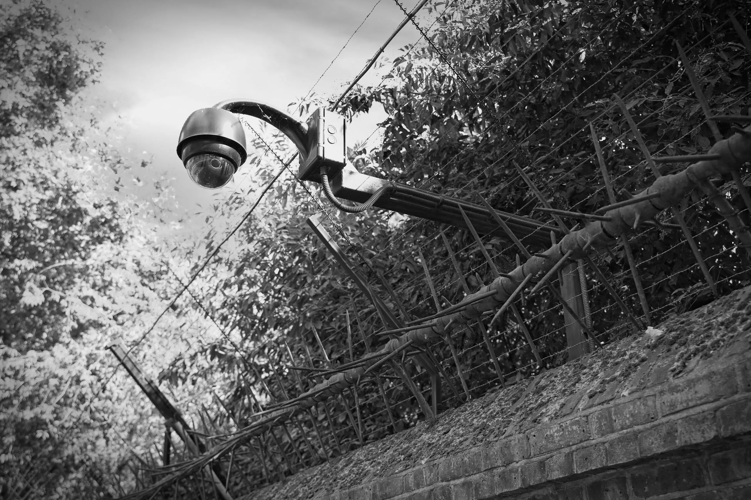 Black and white photograph of a dome security camera mounted above a brick wall topped with barbed wire and metal spikes, England.