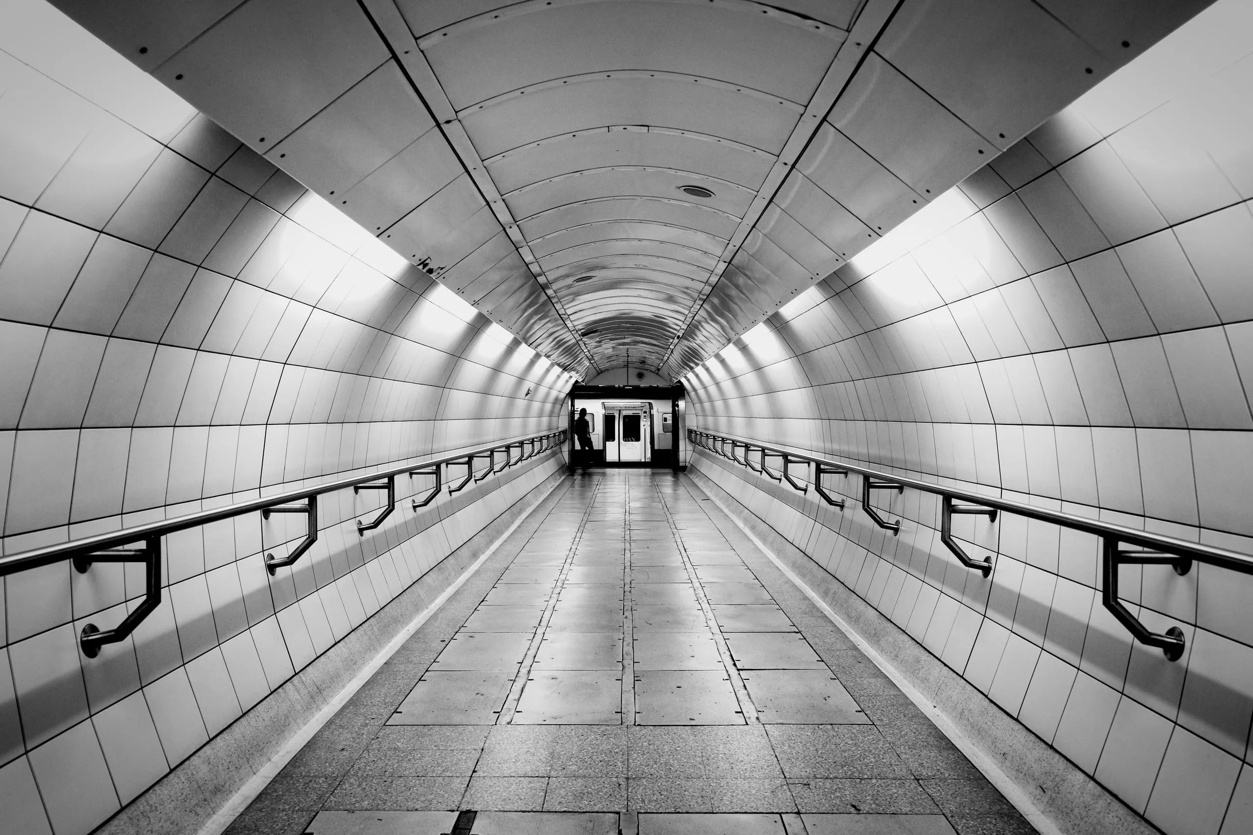Black and white photograph of a symmetrical, white-tiled London Underground passageway with handrails along both walls, leading to a distant doorway where one figure stands silhouetted.