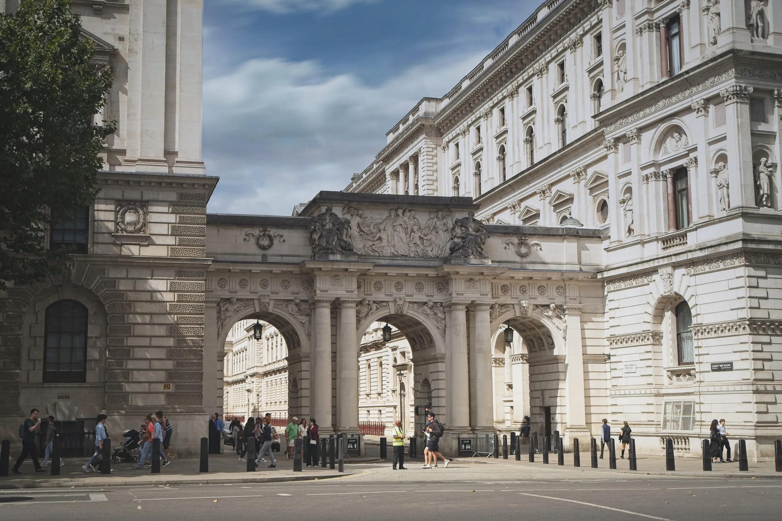 Color photograph of an ornate stone triple-arched gateway spanning a Whitehall street in London, sculpted reliefs above the arches, pedestrians passing beneath near a Cabinet War Rooms sign.