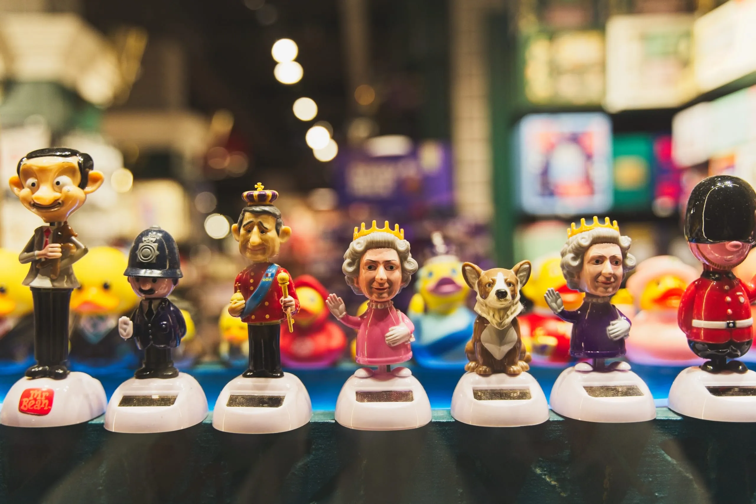 Photograph of a lineup of solar-powered souvenir bobblehead figurines including Mr. Bean, a police officer, royalty, and a palace guard, on a market stall shelf in London, England.