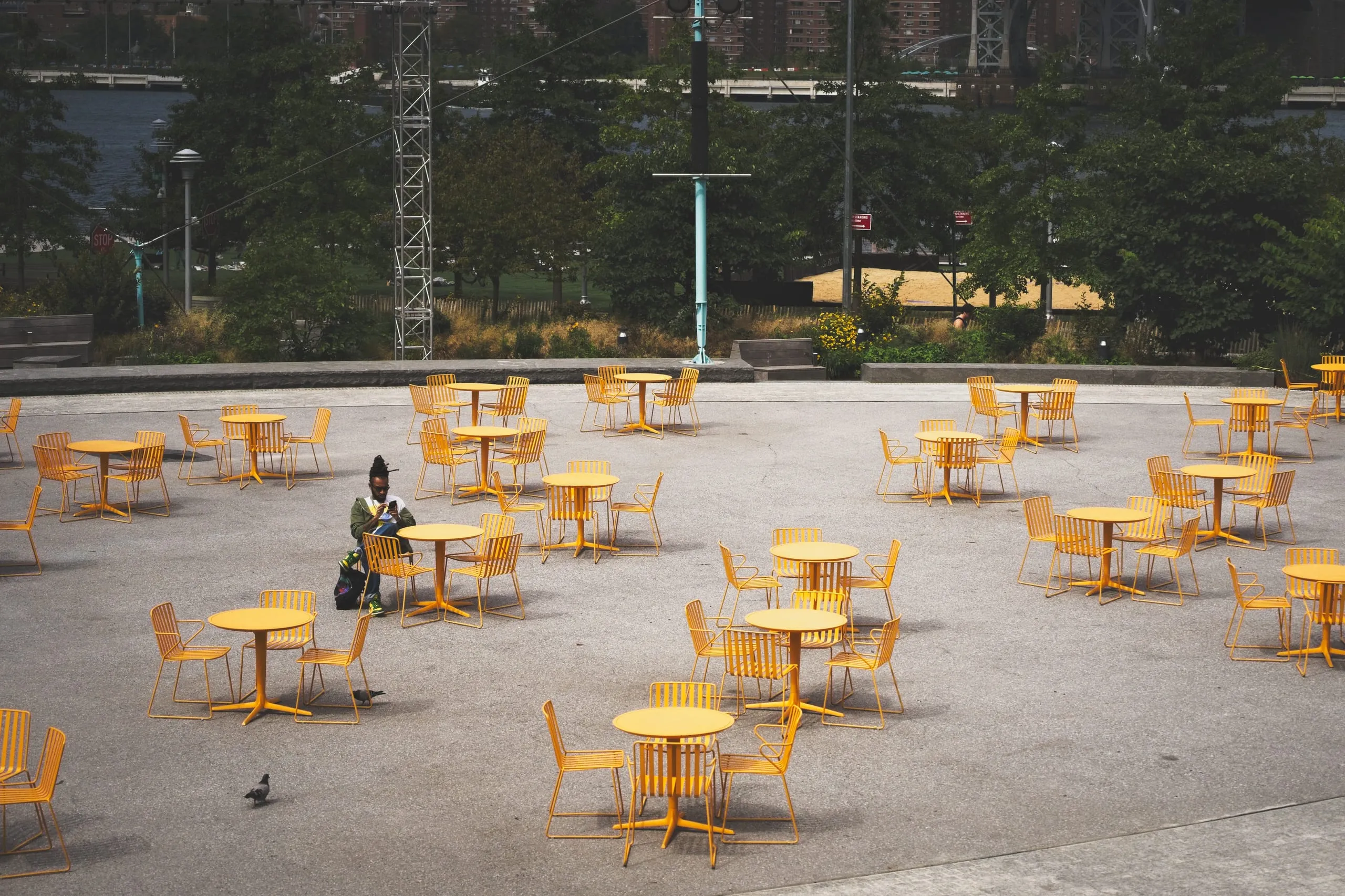 A color photograph of an empty outdoor plaza in Brooklyn filled with rows of bright yellow tables and chairs, a lone person seated among them, and the Williamsburg Bridge visible in the distance.