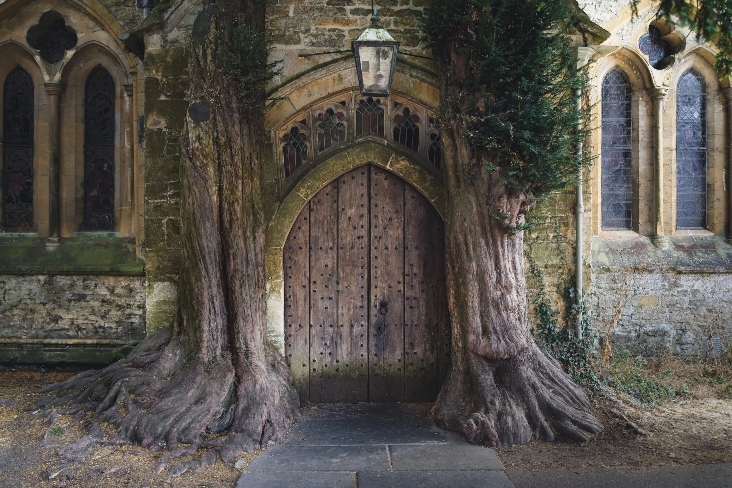A photograph of the ancient wooden door of St Edward's Church in Stow-on-the-Wold, the Cotswolds, England, flanked by two massive old yew trees growing against the stone archway.