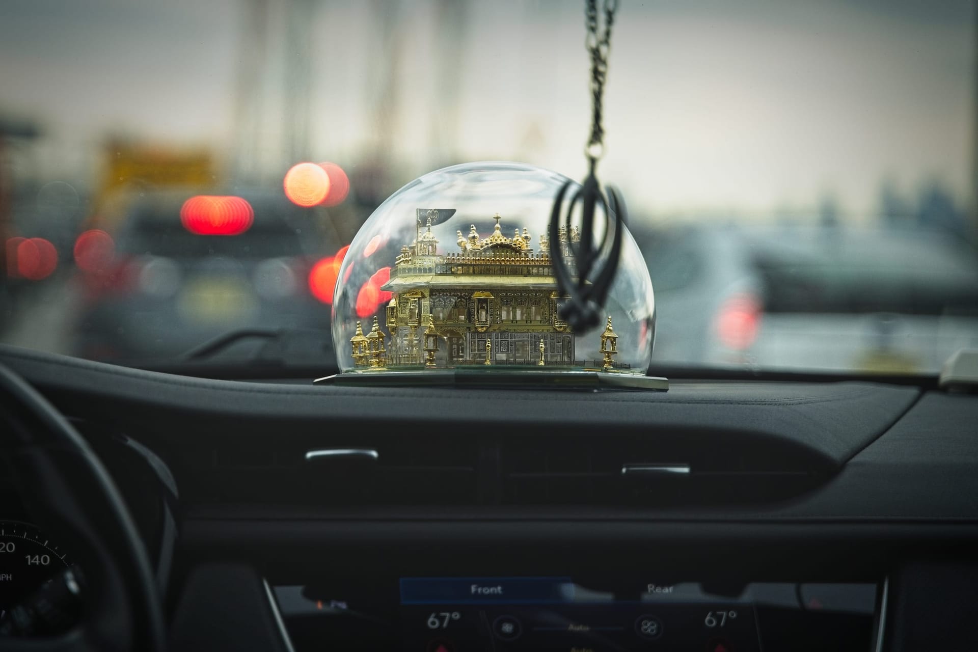 A color photograph of a small golden temple ornament under a glass dome on a car dashboard, with blurred red taillights of Brooklyn traffic visible through the windshield at dusk.