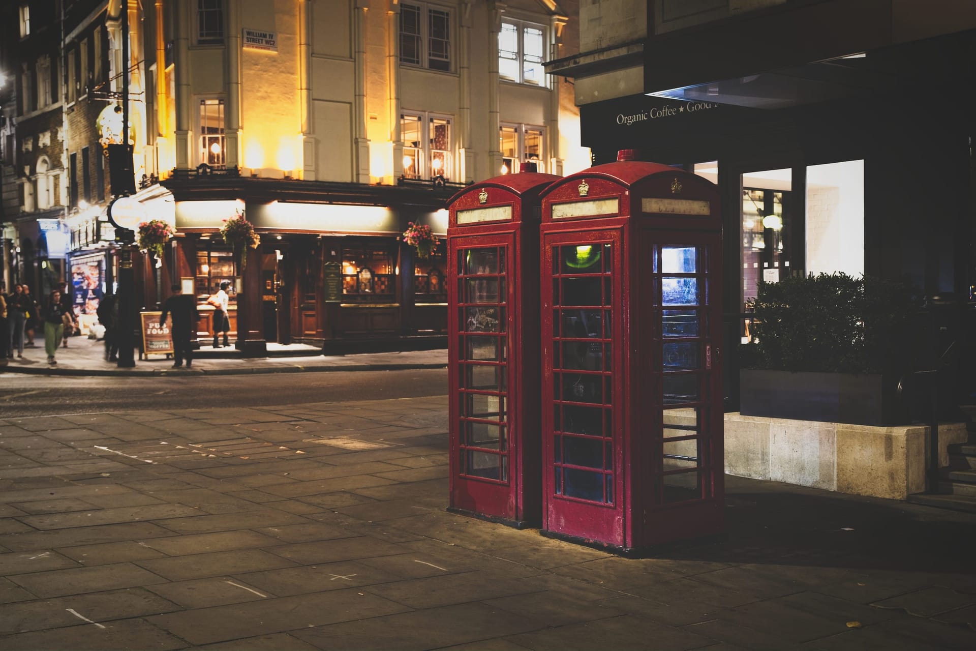 A night photograph of two classic red telephone boxes on a London street corner near William IV Street, with a warmly lit pub in the background.